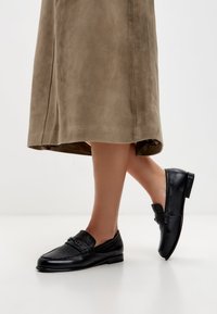 Černé kožené mokasíny s kulatou špičkou, předním páskem a jemnými texturovými detaily. Nohou s taupe sukní, ukazující elegantní design.