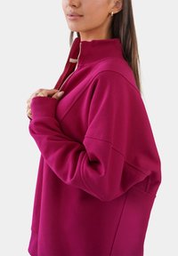 Sudadera de color fucsia con cuello alto, diseño de media cremallera, hombros caídos y textura suave, con bolsillos laterales.