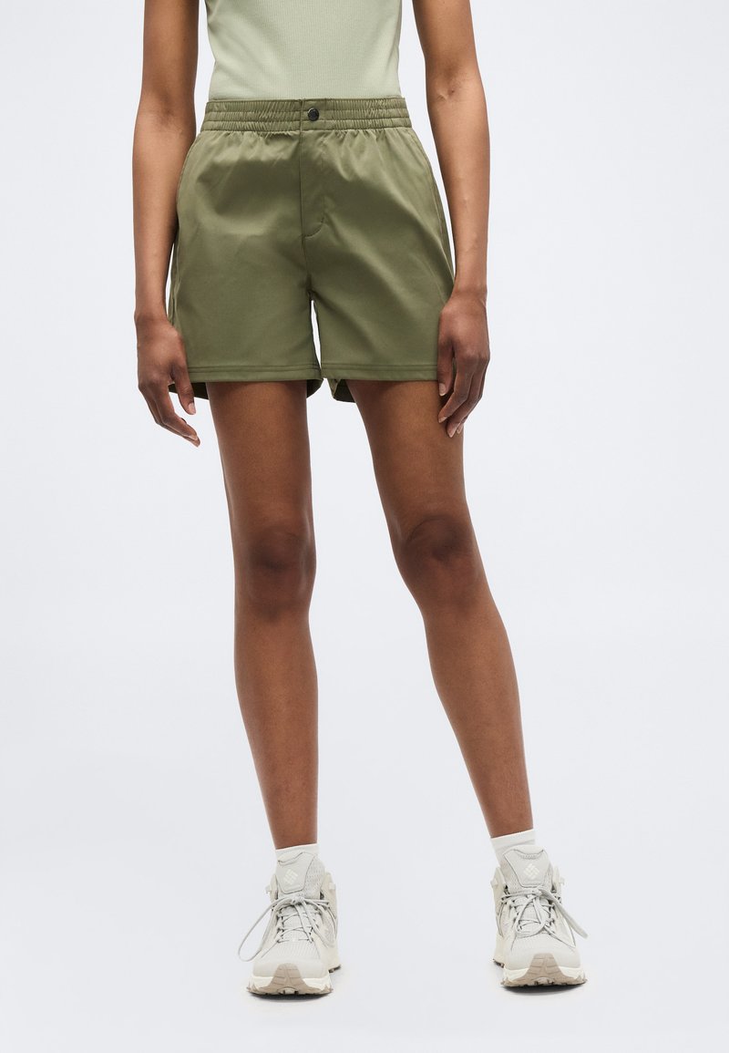 Άτομο που φοράει χακί σορτς με ελαστική μέση, ανοιχτόχρωμο φανελάκι, λευκές κάλτσες και μπεζ αθλητικά παπούτσια, στέκεται μπροστά σε φωτεινό φόντο.