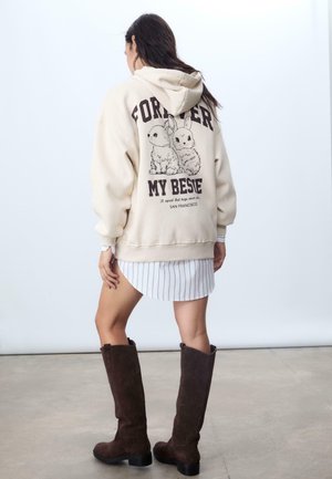 Sweatshirt à capuche crème avec un graphique noir de deux lapins et le texte "FOREVER MY BESTIE" au dos, associé à une robe rayée et des bottes marron.
