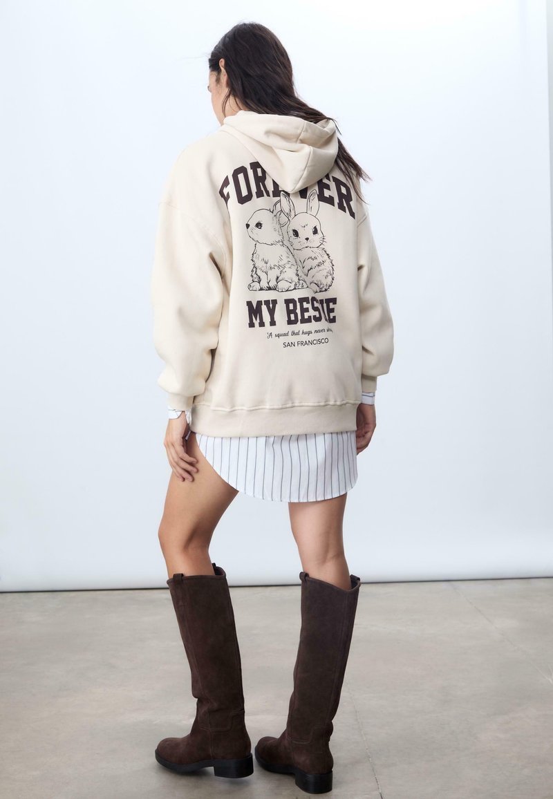 Sweatshirt à capuche crème avec un graphique noir de deux lapins et le texte "FOREVER MY BESTIE" au dos, associé à une robe rayée et des bottes marron.