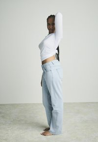 Witte lange mouwen top, aan de zijkant geknoopt, gecombineerd met lichtblauwe rechte jeans, tegen een neutrale achtergrond. Blote tenen zichtbaar.