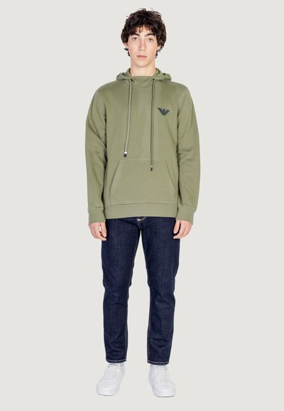 Sweat à capuche vert olive avec cordons, poche kangourou et détail de logo noir sur la poitrine, associé à un jean foncé et des baskets blanches.