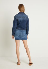 Giacca di jeans di colore blu scuro, presenta un design classico con una forma aderente. Abbinata a una mini gonna di jeans coordinata, entrambi i capi hanno delle tasche.