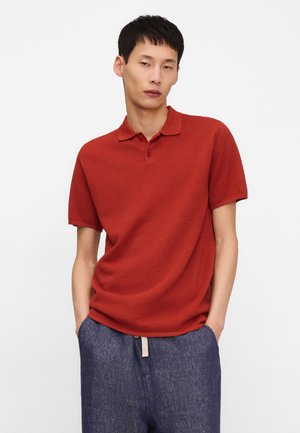 Mężczyzna w czerwonej koszulce polo z krótkim rękawem i luźnych niebieskich spodniach, stojący z rękami w kieszeniach na jednolitym tle.