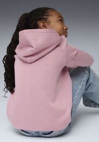 Helle rosa Kapuzenjacke mit Kordelzug, aus weichem Stoff. Getragen mit ausgewaschenen blauen Jeans, in sitzender Position, mit strukturiertem Haar.