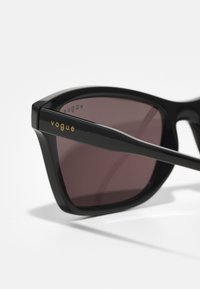 Óculos de sol pretos com um design geométrico, apresentando lentes escuras e a marcação "vogue" em dourado na haste. Textura de plástico lisa.