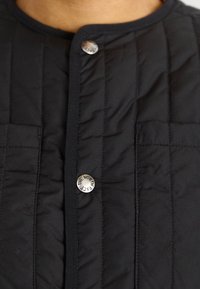 Black quilted jacket featuring a round neckline, vertical seam detailing, and a metallic snap closure with logo accent.