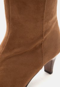 Gros plan d'une bottine en daim marron avec des coutures visibles et un petit talon en bois empilé sur fond blanc.