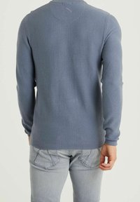 En man som bär en långärmad blå texturerad tröja och ljusgrå jeans, står med ryggen mot kameran och har höger hand avslappnat vid sidan.