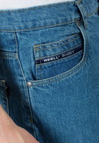 Jeans holgados de color azul claro con una tela texturizada, que presentan costuras en contraste, un bolsillo trasero con la etiqueta "REELL BAGGY" y un botón de metal.