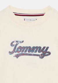 Sweatshirt crème à col rond, arborant un logo "Tommy" en sequins bleus accentué de points multicolores et de détails rayés à l'intérieur du col.