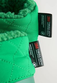 Bota verde, resistente al agua, con diseño acolchado y forro de peluche. Las etiquetas indican aislamiento 3M Thinsulate; detalles del producto mostrados de manera destacada.
