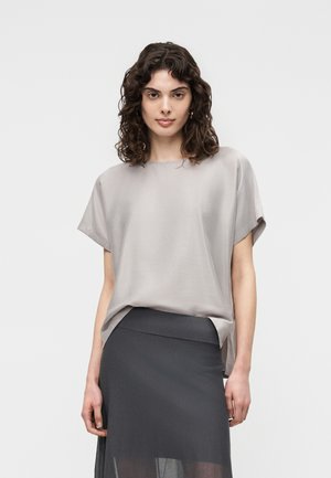 Femeie cu păr creț și închis la culoare poartă un top lejer gri deschis și o fustă gri închis cu talie înaltă, pe un fundal simplu, deschis la culoare.