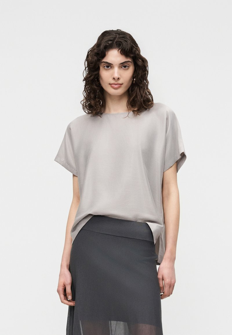 Femme aux cheveux bouclés et foncés portant un haut ample gris clair et une jupe taille haute gris foncé sur un fond clair uni.