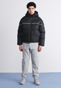 Sort pufferjakke med hætte, med en hvid vandret stribe, parret med lysegrå sweatpants og grå sneakers.