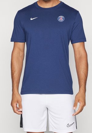 Homme portant un t-shirt bleu marine du Paris Saint-Germain et un short blanc Nike, debout les bras détendus le long du corps.