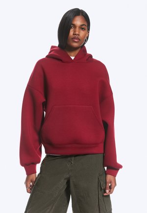 Femme portant un sweat à capuche rouge ample avec une poche avant et un pantalon cargo vert olive, debout devant un fond blanc uni.