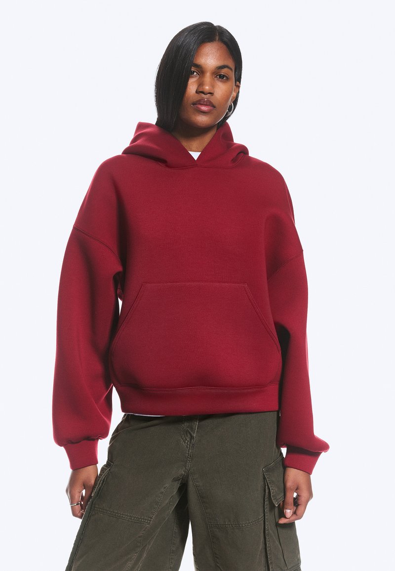 Femme portant un sweat à capuche rouge ample avec une poche avant et un pantalon cargo vert olive, debout devant un fond blanc uni.