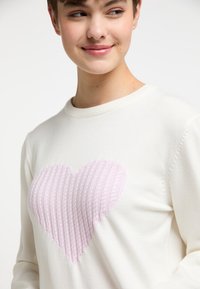 Un pull créme avec un motif cœur en tricot texturé de couleur rose clair sur la poitrine, un col rond et des poignets côtelés.
