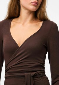 Sieviete valkā pieguļošu tumši brūnu apvalka kleitu ar garām piedurknēm un V-veida dekoltē, kas sasieta jostasvietā ar atbilstošu jostu.