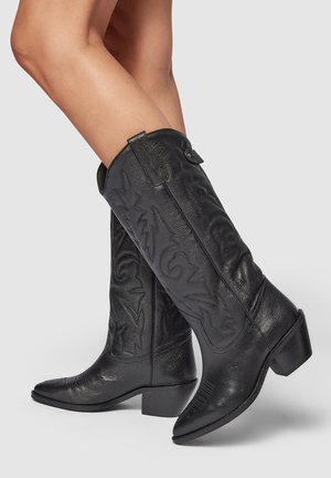 Bottes de cowboy en cuir noir avec des coutures décoratives surélevées, un embout pointu, un talon bas empilé et un bouton d'accentuation à l'arrière supérieur.