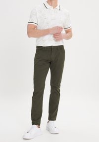 Witte polo met bloemendessin, donkergroene broek en witte sneakers. Het shirt heeft een kraag met donkere afwerking en korte mouwen.