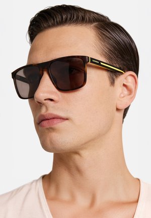 Giovane uomo con capelli pettinati all'indietro che indossa occhiali da sole marroni tartarugati con strisce gialle e lenti scure, rivolto verso sinistra.