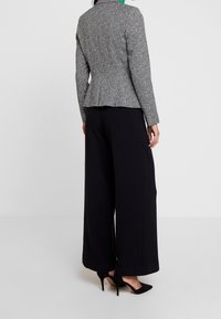 Blazer gris à motif chevrons avec taille péplum, associé à un pantalon noir à jambes larges et des escarpins noirs à bouts pointus. Design minimaliste.