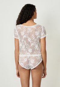Top in pizzo bianco con maniche corte, motivo floreale e bordi ondulati, abbinato a pantaloni in pizzo coordinati. Tessuto trasparente e leggero.
