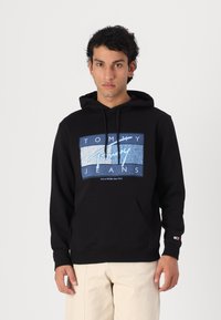 Hoodie noir avec un design graphique aux éléments bleus et blancs. Comprend une poche avant et un capuchon avec cordon de serrage. Coupe classique.