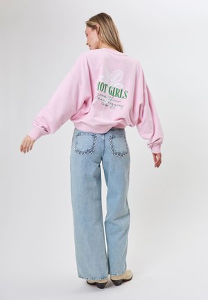 Frau trägt einen pinken oversized Pullover mit dem Schriftzug „HOT GIRLS“ auf dem Rücken und hellblaue Jeans mit gewellten Taschenkanten, steht vor weißem Hintergrund.