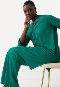 Grön, långärmad, lös passande tunika med nyckelhålsutskärning, matchad med vida byxor. Slät tyg med mjuk textur och minimala designdetaljer.