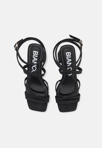 Musta hihnallinen korkokenkä, jossa on nilkkasolki, neliön muotoiset varpaat, brändätty valkoinen teksti pohjallisiin, nähtynä ylhäältä valkoisella taustalla.