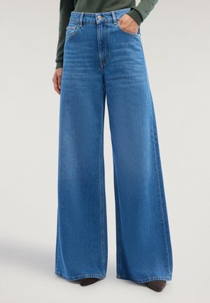 Personne portant un jean bleu taille haute à jambes larges, une main dans une poche, associé à des chaussures pointues foncées et un haut vert à manches longues.