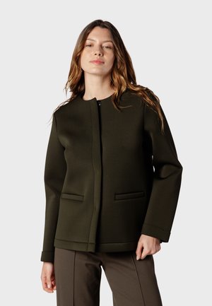 Femme aux longs cheveux portant une veste vert foncé sans col et un pantalon marron, debout devant un fond clair uni.