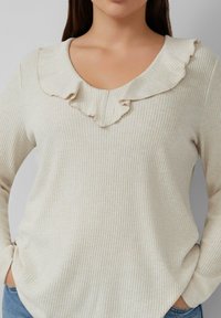 Helles beige, geripptes Oberteil mit V-Ausschnitt und Rüschensaum. Lange Ärmel und lockere Passform, getragen mit lässigen blauen Jeans.