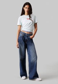 Lichtblauwe cropped t-shirt met een rood logo, gecombineerd met oversized donkerblauwe denim jeans. Model draagt witte sneakers, met handen in de zakken.