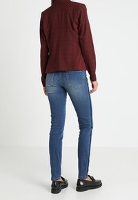 Žena v úzkých modrých džínách s bočními pruhy, v vínově červeném kostkovaném saku a černých mokasínech, stojící na bílém pozadí.