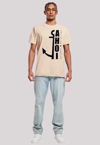 Beige Baumwoll-T-Shirt mit auffälligen schwarzen Grafiken, Text und einem Ankerdesign. Kombiniert mit hellblauen Jeans und weißen Turnschuhen.