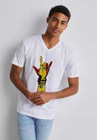 Camiseta blanca de cuello en V con un gráfico de una mano amarilla con acentos rojos y detalles negros, sobre un fondo liso.