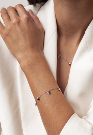 Arm with silver bracelet featuring small heart charms, resting on opposite shoulder, wearing white blazer.
