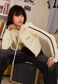 Kremasta flis bunda s prednjim zavrsnicom, crnim zvjezdastim akcentima i prugama duž rukava. Model nosi tamnosive široke traperice.