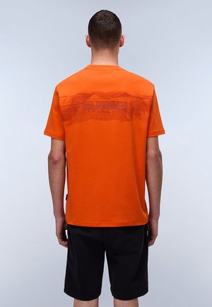 Oranžové bavlněné tričko s krátkým rukávem, na zadní straně s texturovaným grafickým designem, kombinované s černými šortkami.
