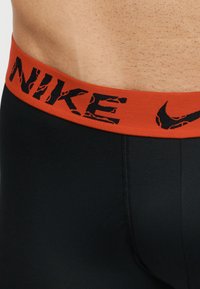 Μέση και κάτω μέρος κορμού με μαύρο σορτς με πορτοκαλί μέση που έχει μαύρο φθαρμένο λογότυπο Nike και σχέδιο swoosh.