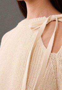 Kremowy sweter dzianinowy z odkrytymi ramionami, z wstążką przy dekolcie. Tkany materiał z prążkowanym wzorem.