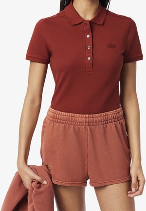 Mujer con una camisa polo color óxido y pantalones cortos con cintura elástica a juego, sosteniendo una sudadera doblada en una mano.