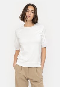 White knitted top with short raglan sleeves, crew neck, and ribbed hem. Fabric appears soft with a smooth texture. Model poses with hands in pockets.