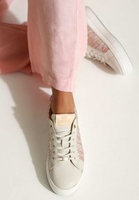 Beżowe sneakersy z plecionymi różowo-fioletowymi panelami bocznymi, białą gumową podeszwą i brązowym logo na pięcie. Stylizowane z jasnoróżowymi spodniami.