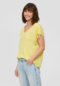 Gelbes V-Ausschnitt-T-Shirt aus weichem Stoff, mit kurzen Ärmeln und Bindedetail sowie lockerem Schnitt, kombiniert mit hellblauen Jeans.